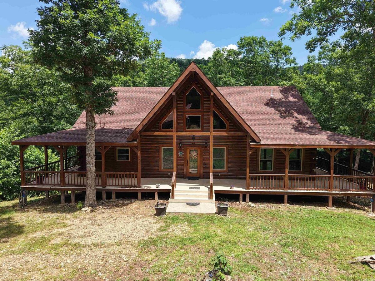 Front of Sugar Creek Cabin: timber-frame gable, log walls and the wraparound porch under the oaks